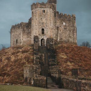 Vertical photograph of the medieval Norman keep at Cardiff Castle atop a earth mound with a Welsh flag flying against a dark sky