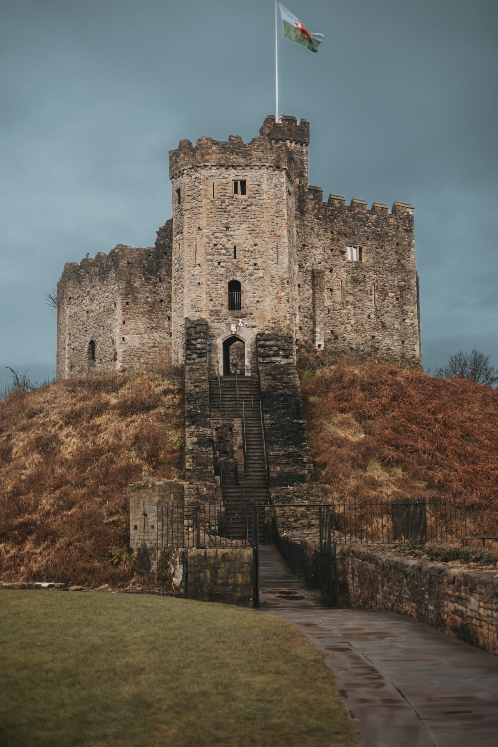 Vertical photograph of the medieval Norman keep at Cardiff Castle atop a earth mound with a Welsh flag flying against a dark sky