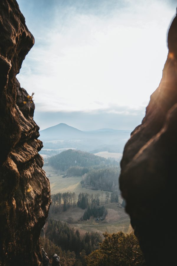 A distant mountain and rolling Czech landscape viewed through a narrow opening between towering sandstone rock formations.