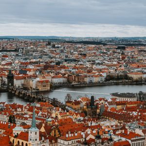 High-angle panoramic view of Prague displaying red terracotta rooftops, Charles Bridge, and the Vltava River under a cloudy sky