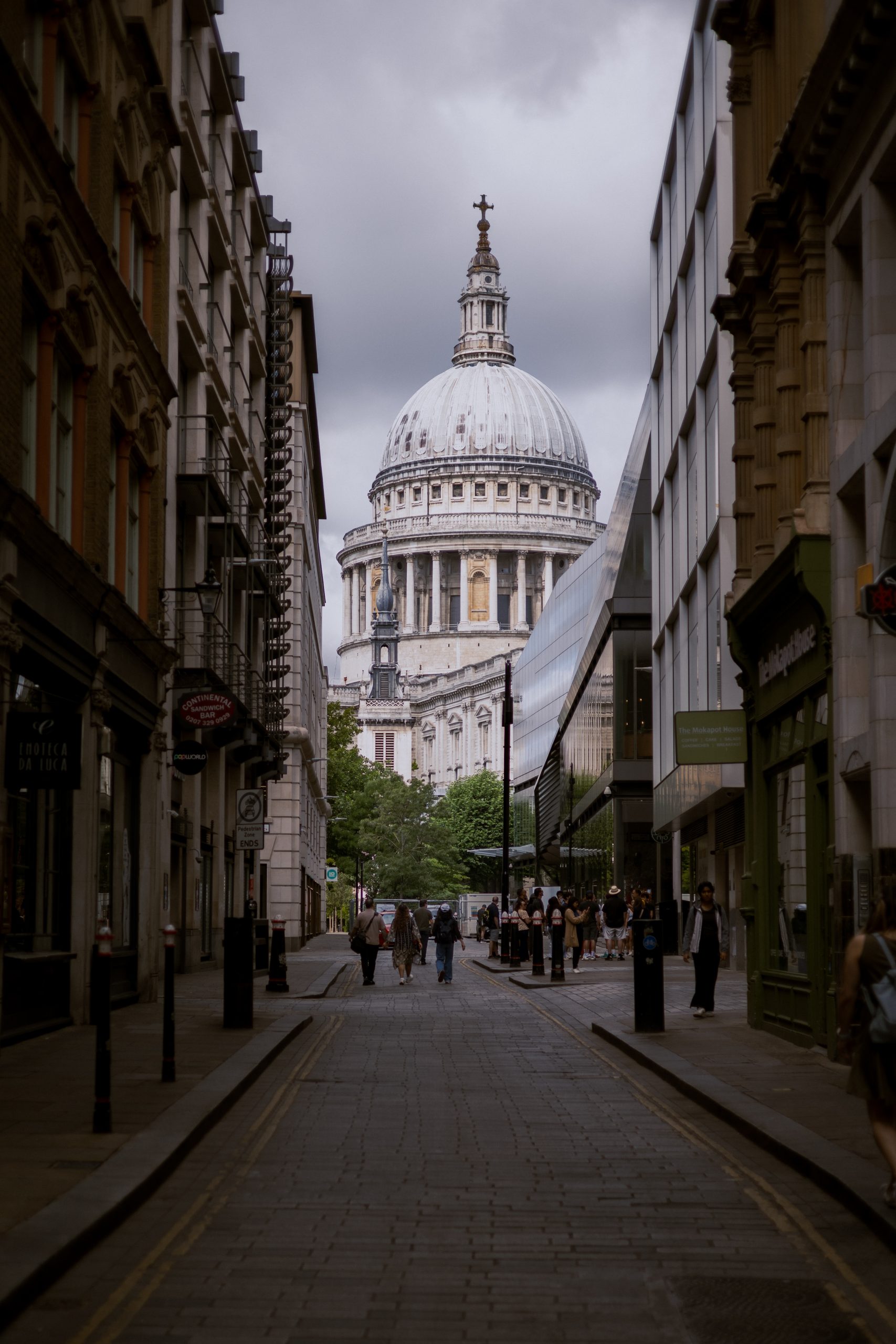 View looking down a cobblestone London street framed by historic and modern buildings leading to St Paul's Cathedral dome under an overcast sky