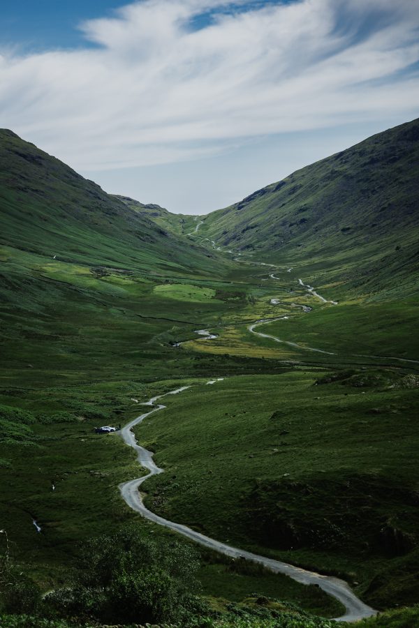 A winding single track road leads through a green Lake District valley, following a meandering stream between steep hills beneath a sweeping cloudy sky.