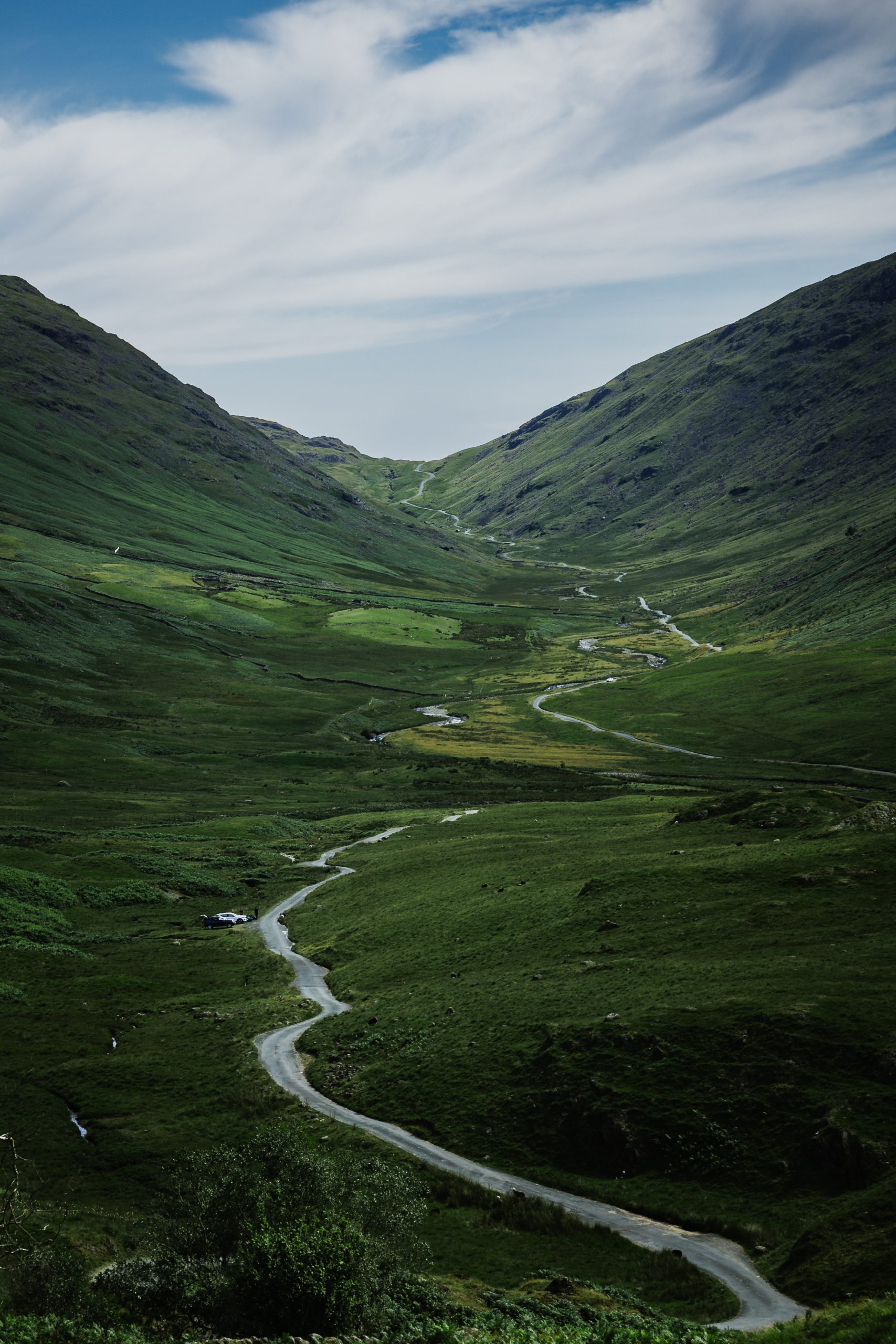 A winding single track road leads through a green Lake District valley, following a meandering stream between steep hills beneath a sweeping cloudy sky.