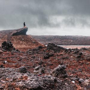 Landscape photograph of a lone silhouette standing atop a jagged volcanic crater ridge in Iceland under an overcast sky